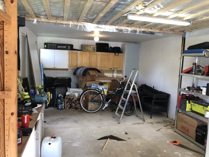 Ett halvtomt garage med öppen takstol, diverse verktyg, en stege, en cykel och gamla köksskåp längs väggen.