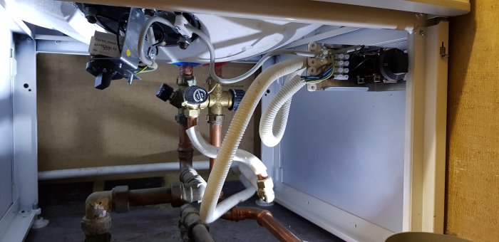 Undersida av en värmepanna med röranslutningar och elektriska komponenter vid en vägg.