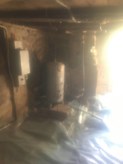 En suddig källare med stenväggar, träbjälkar, en varmvattenberedare och skyddsplast över golvet.