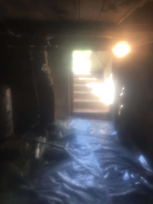 Källare med ståhöjd under hus, jordstampat golv med plast, stentrappan uppåt och belysta trätakbjälkar.