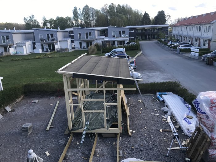 Byggplats med oavslutat takprojekt och torra brädor säkrade på hustaket, med radhus i bakgrunden.