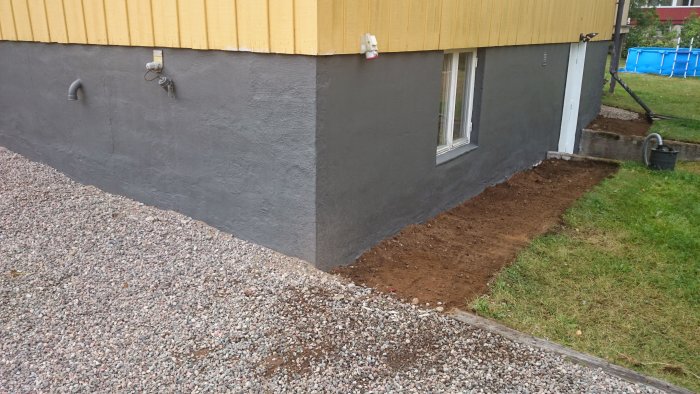 Nymålad grå sockel på hus, uppluckrad jord längs väggen, grus och gräsmatta.