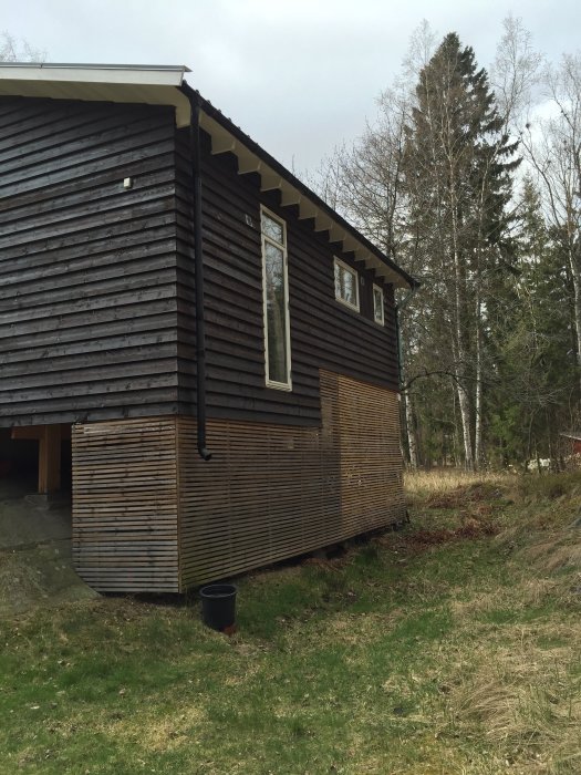 Friliggande hus med mörkt träpanel och spaljéer på altanerna, nära skog utan synliga grannar.