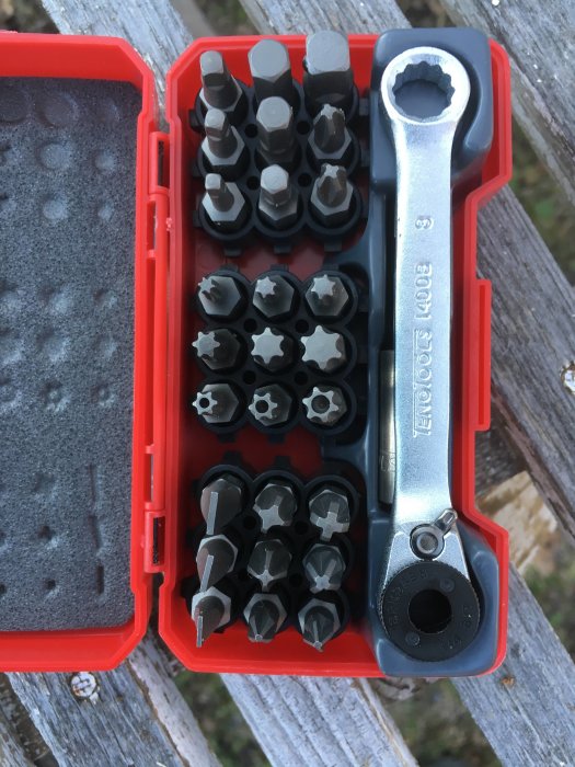 Öppen ask med olika storlekar av bits för skruvdragare bredvid en ringnyckel på ett träunderlag.