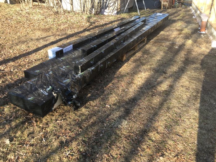 Inplastade limträbalkar och pelare staplade utomhus, nära en byggarbetsplats.