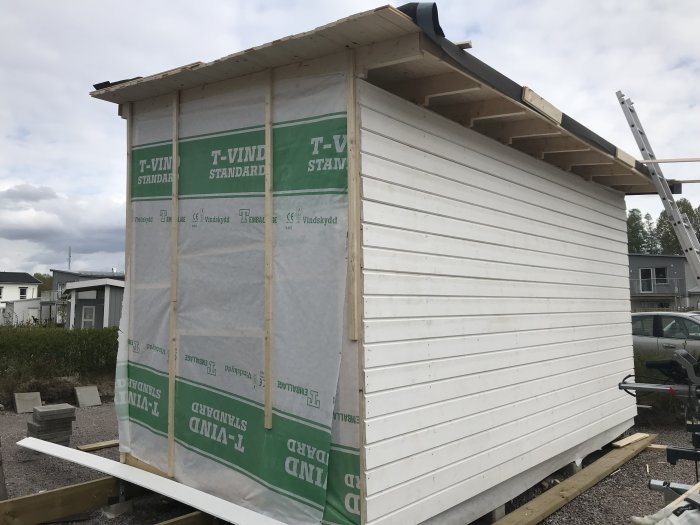 Delvis klädd husvägg med träpanel och väderskyddande plastfolie, byggmaterial i förgrunden.