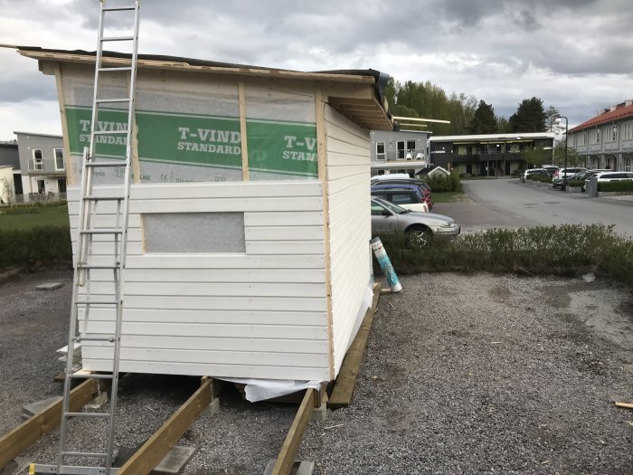 Hus under renovering med delvis monterad vit träpanel och en stege, på en byggplats med bilar i bakgrunden.