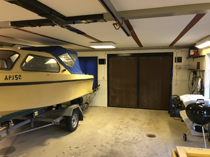 Inredning av ett uthus med båt på trailer, betonggolv och bruna dubbla garageportar.