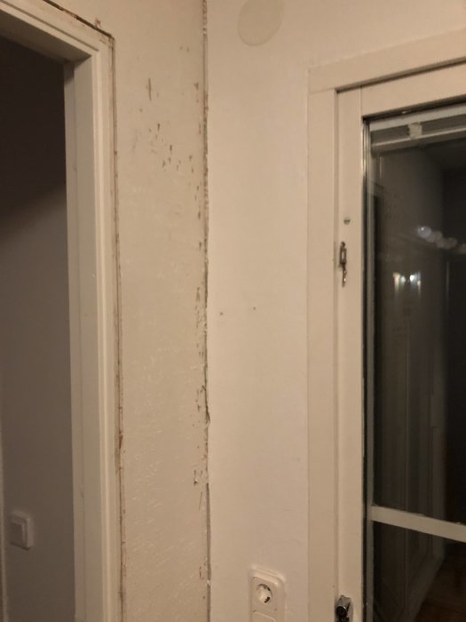Skadad och flagnande vitmålad vägg vid en dörrkarm och ett eluttag, behöver reparation.
