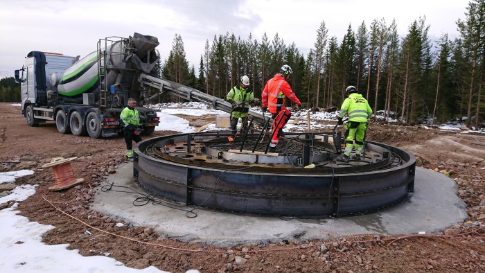 Arbetare gjuter betong vid en vindturbinfundament med betongbil och vibrationsverktyg i skogsområde.