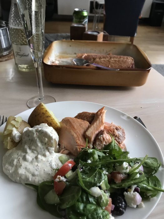 Tallrik med rökt lax, potatis, sås och sallad, bakplåt och ett glas vin i bakgrunden.