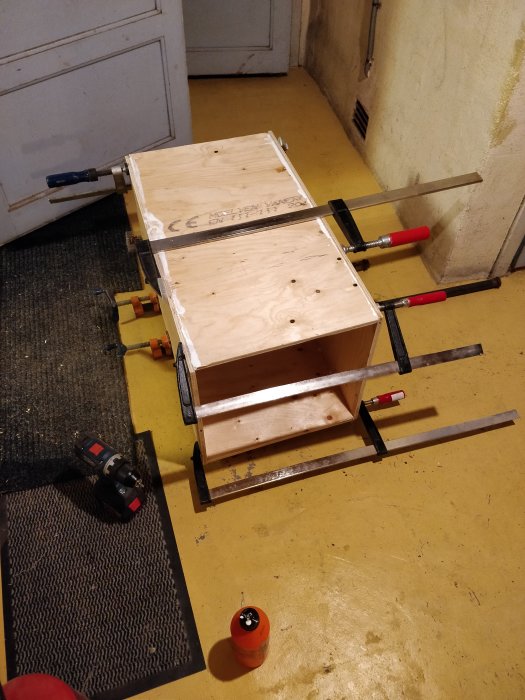 Träbyggnadsprojekt i fastsättningsfas med klämmor och en borrmaskin på ett verkstadsgolv.