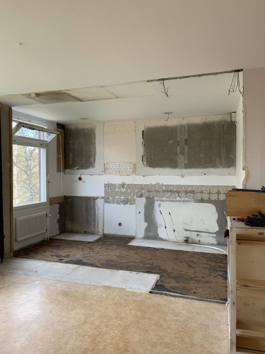 Kök under renovering med delvis borttaget golv och väggbeklädnad, exponerad betong och rördragningar.
