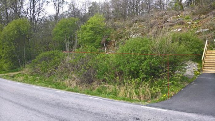 Vy av ett sluttande grönområde intill en väg, markerat för höjning med gabioner planerade längs kanten.