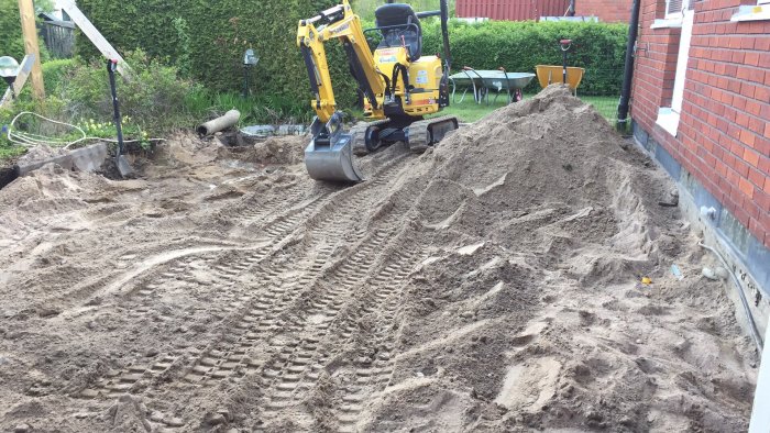 Grävmaskin i arbete på en tomt med en stor hög av sand och grus, spår i marken indikerar nyligen utfört arbete.