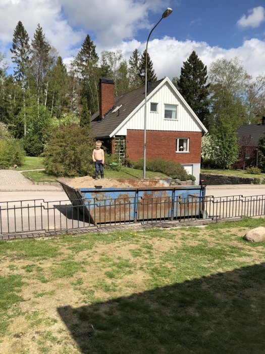 En person står bredvid en blå container full med sand och grus framför ett hus.