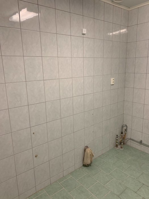 Tomt hörnrum med kaklade väggar och grönaktigt golv som visar vattentillförseln från en tidigare pissoarinstallation.