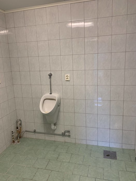Badrum med kaklade väggar och pissoar, synliga rör och vattenanslutningar, planer för renovering.