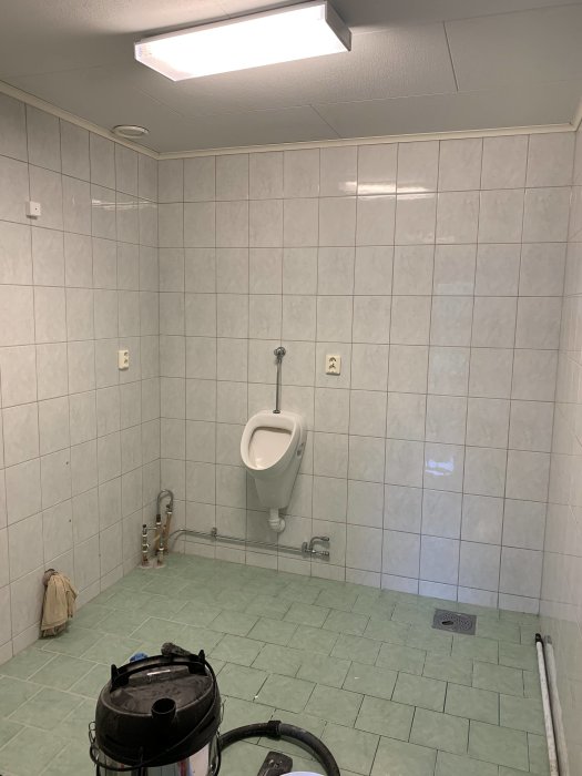 Renoveringsprojekt av ett badrum med pissoar på väggen och vattenrör synliga, före ombyggnad.