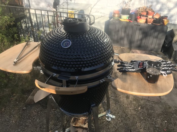 Kamado grill med öppna sidobord, grilltång, kötthandskar och ett glas dryck utomhus.