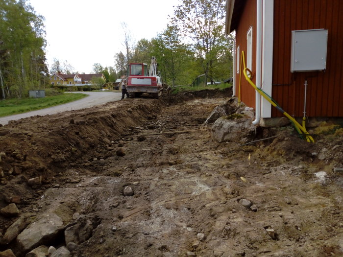 Grävmaskin vid pågående grävningsarbete nära hus för fundament till kantbalk.