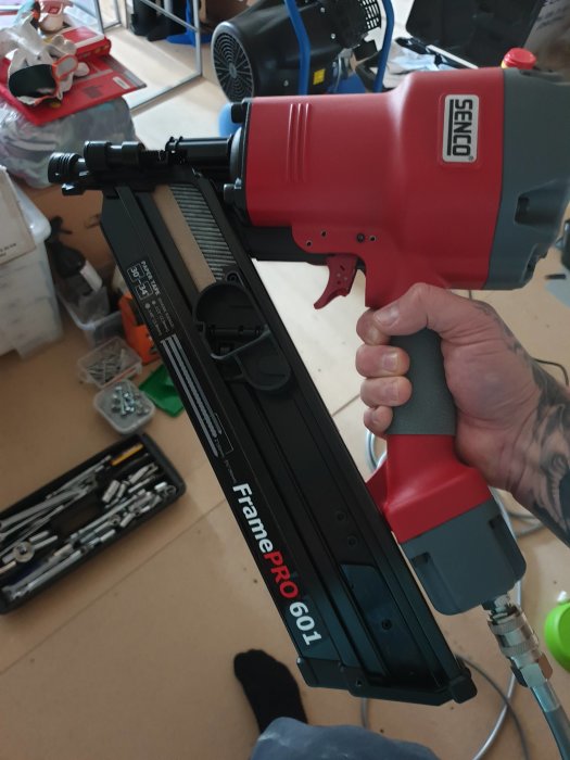 En person håller en röd och grå Senco spikpistol, modell FramePro 601, inomhus.