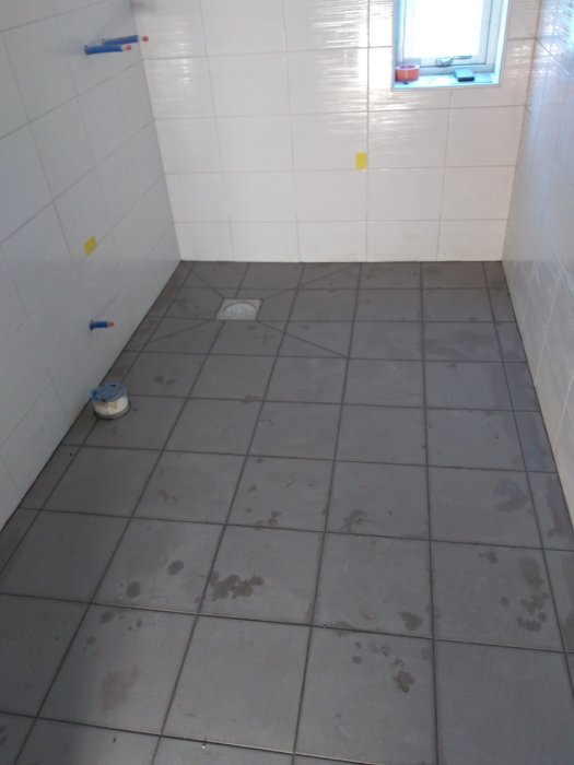Ett nyligen spacklat badrumsgolv med mörka klinkerplattor och vita kaklade väggar, utrustningar ligger synligt.