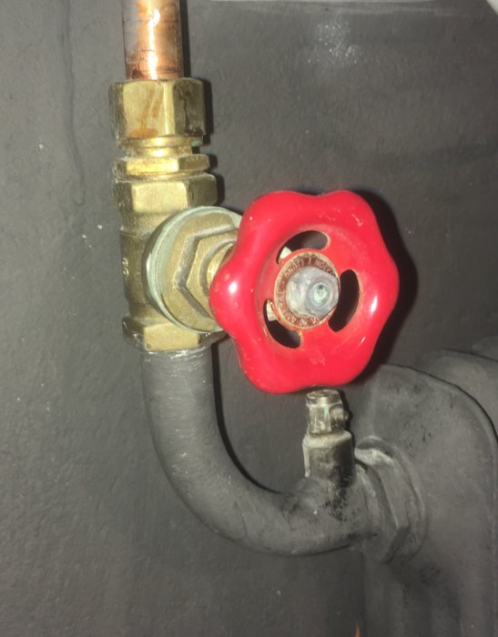 Gammal röd radiatorventil med anslutningsrör i koppar och prisol ovanpå en grå vägg.