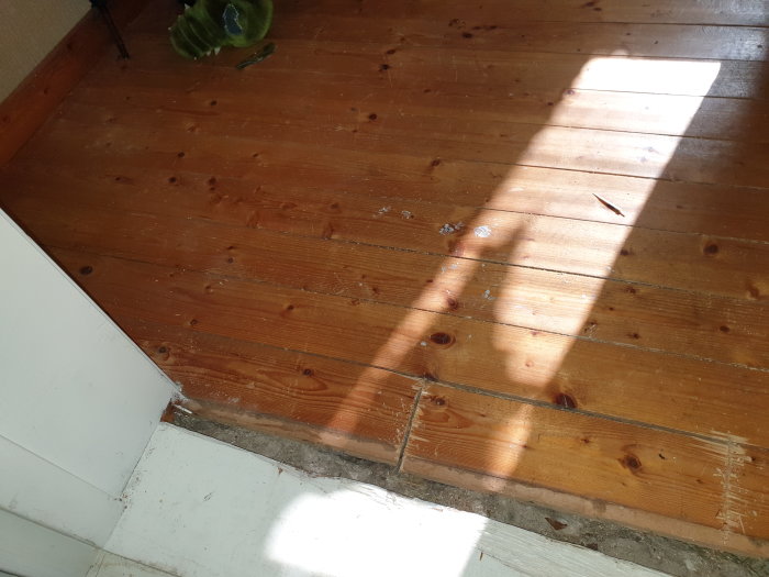 Trägolv i källare med upplyft planka som avslöjar underliggande isolering, solljus genom fönster.