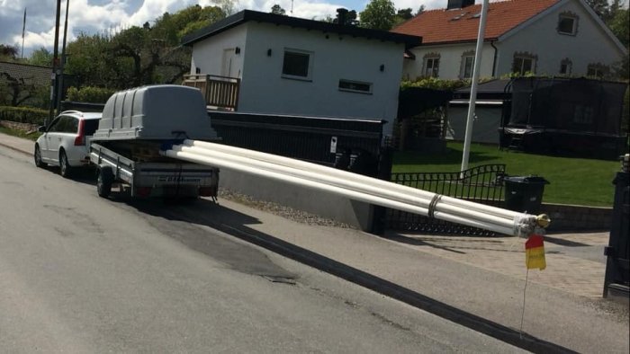 Släpvagn med långa vita rör som sticker ut bakom, säkrade med varningsflagga på en gata.