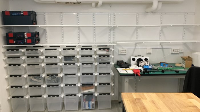 Organiserad förvaringslösning med etiketterade lådor och hyllor monterade med elfas skensystem i ett arbetsrum.