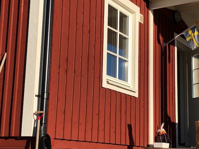 Del av ett hus med faluröd lockläktspanel och vita knutar nära ett fönster, synliga glipor i träet.