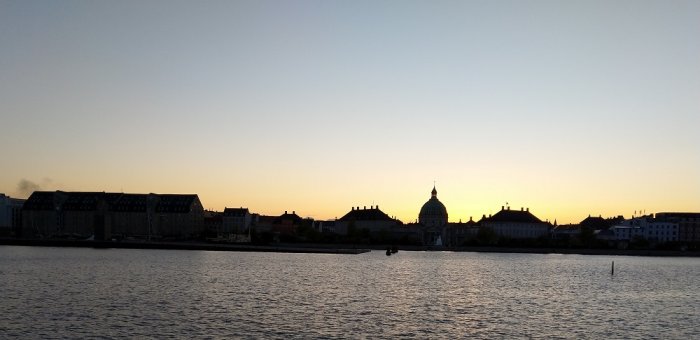 Solnedgång över stadssilhuett med vatten i förgrunden och en klar himmel.