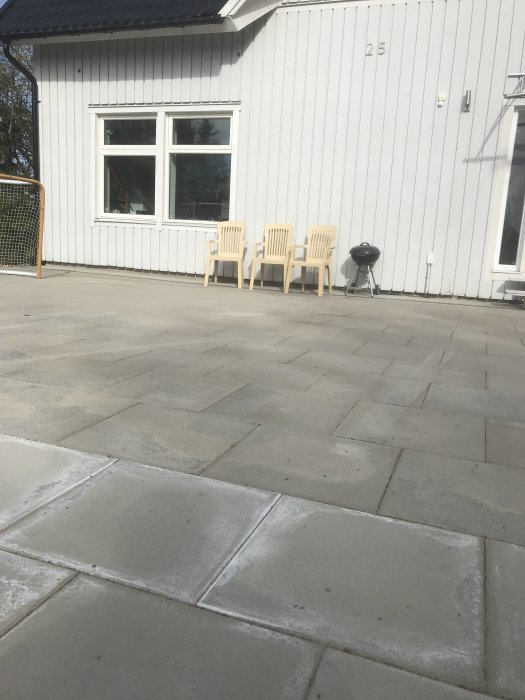 Vit putsad husvägg med två stolar och en liten grill på en stenlagd uteplats, föreslagen plats för matbord.