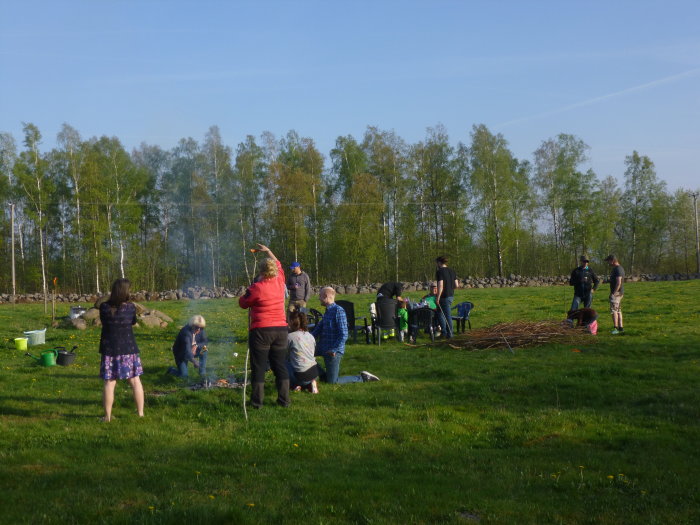 Grupp människor samlade utomhus runt en liten brasa, vissa grillar korv och umgås på en grön äng.