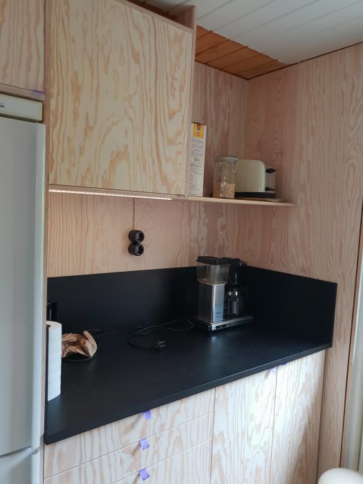 Kökshörna i fritidshus med oslipad plywood på väggar och skåpluckor samt svart bänkskiva med köksutrustning.