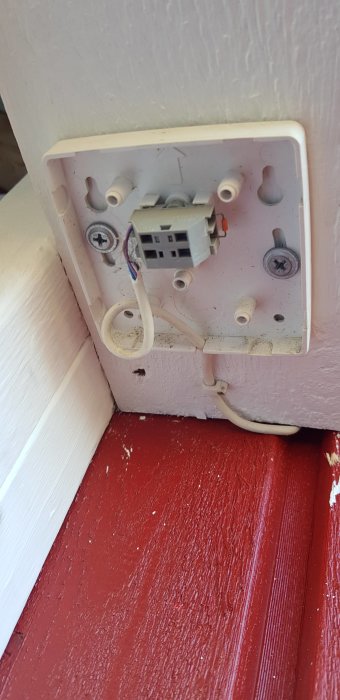 Öppet eluttag utanpå vägg med synliga trådar och installationskomponenter, vid sidan av rödmålat träverk.