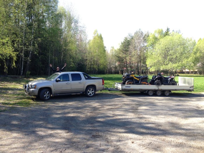 Pickup med trefjädrat släp lastat med fyrhjulingar, person gestikulerar med armarna i bakgrunden.