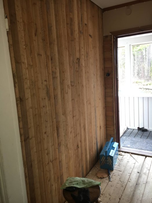 Entréhall med stående träpanelväggar utan synliga spikar, takbalkar ovanför och ett fönster som visar ett skogligt landskap.