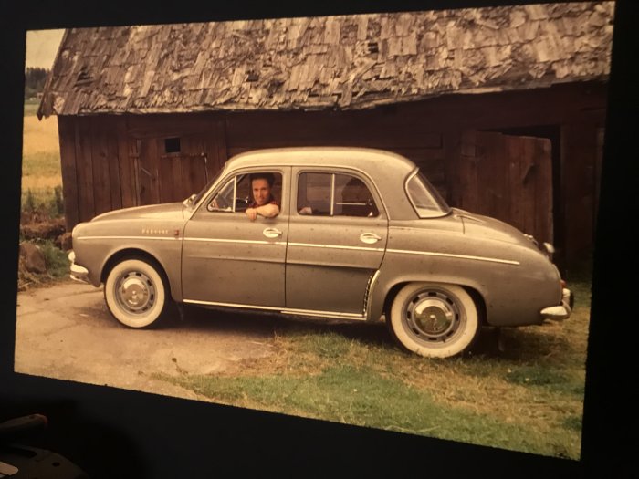 Man som tittar ut från förarsätet i en klassisk Peugeot, parkerad framför lada.