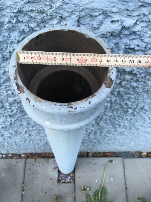 Måttband visar innerdiameter på gammalt gjutjärnsrör mot grå fasad.