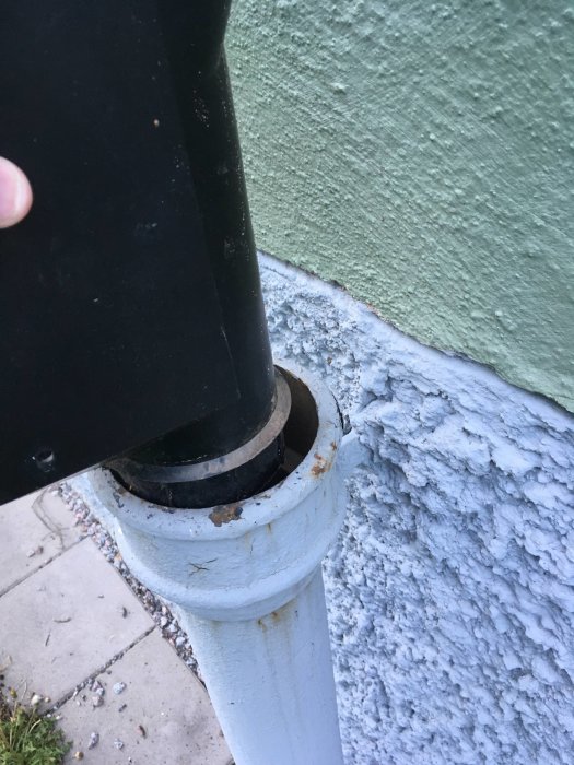 En svart lövsil hålls ovanför ett vitt gjutjärnsrör mot en vägg, lövsilen verkar vara mindre i diameter än röret.