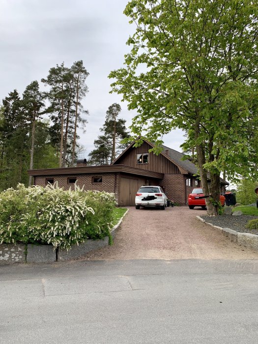 Ett brunt hus från 1970 med träpanel och tegeltak omgivet av grönska och två parkerade bilar i uppfarten.