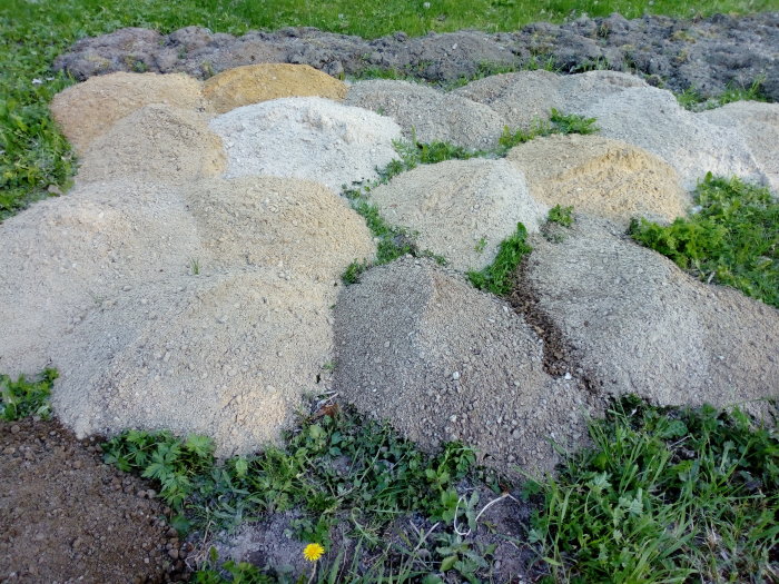 Flera högar av silad sand i olika färger och kornstorlekar utspridda på gräs.