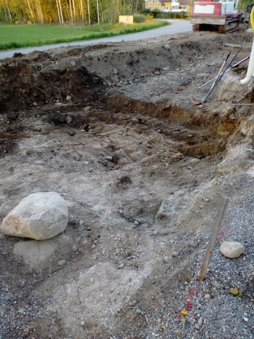 Grävd grop med ojämn sand och jord, stenkil och verktyg synliga, i ett bostadsområde med grönska.