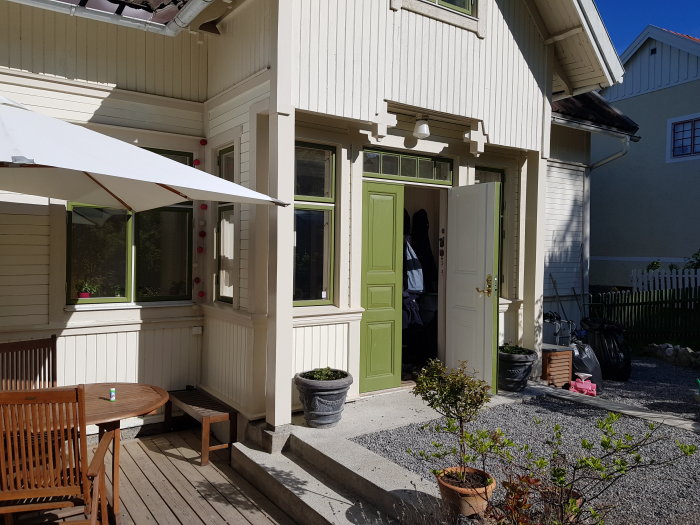 Slutgiltigt renoverat hus med öppen grön ytterdörr och en träterrass med möbler och planteringar i soligt väder.
