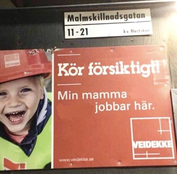 Skylt med texten "Kör försiktigt! Min mamma jobbar här." och leende barn i bygghjälm.