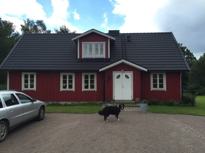 Renoverat rött hus med svart tak, två kupor och en veranda, hund på gården.