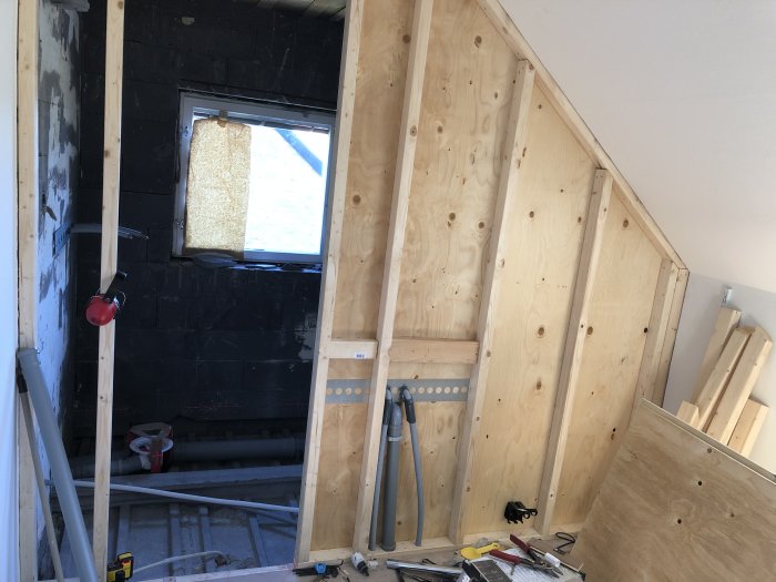 Pågående renovering av ett litet badrum under snedtak med exponerad isolering, väggstommar och VVS-installationer.