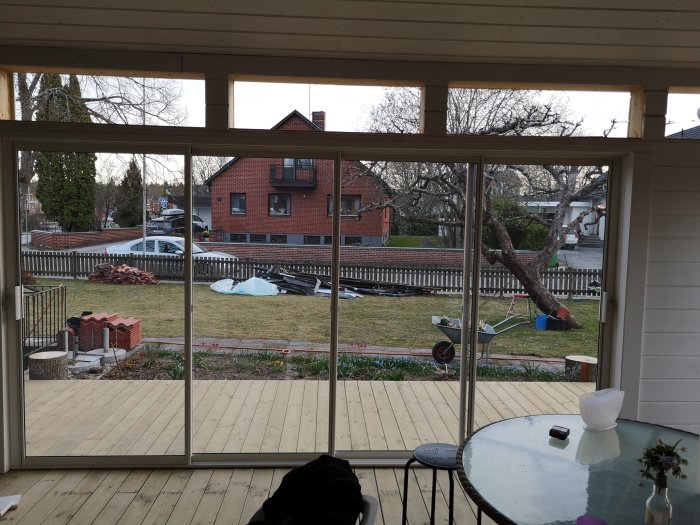 Utsikt från en veranda med nyinstallerade glaspartier ser ut mot en trädgård och ett rött tegelhus.